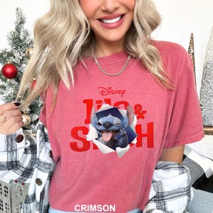 May include: Crimson-colored t-shirt featuring the Disney logo and the words "Lilo & Stitch" in red. The shirt has a graphic of Stitch popping out of a torn paper effect, with his tongue out and a peace sign.