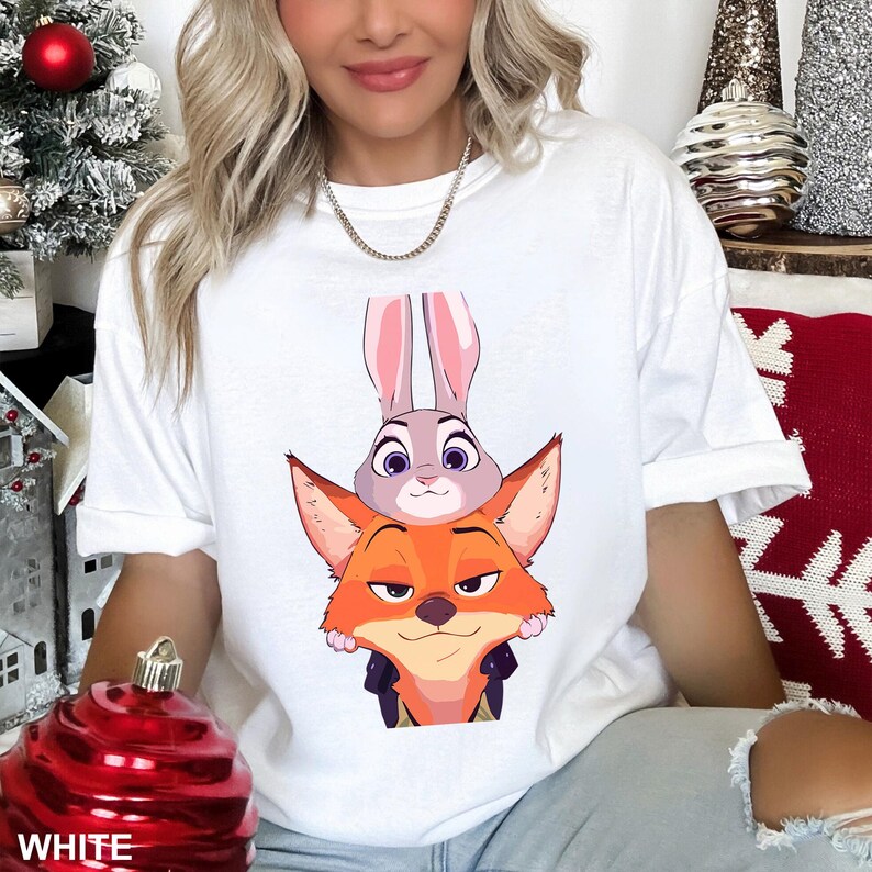 Puede incluir: Camiseta blanca con una ilustraci&oacute;n de dibujos animados de Judy Hopps y Nick Wilde de Zootr&oacute;polis. Judy, una coneja gris, est&aacute; encima de Nick, un zorro naranja. La palabra "WHITE" est&aacute; impresa en la parte inferior de la imagen.