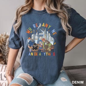 May include: A denim blue t-shirt featuring a colorful graphic of various cartoon characters in front of a cityscape. The text "WHERE ANYONE CAN BE ANYTHING" is displayed in a rainbow font.
