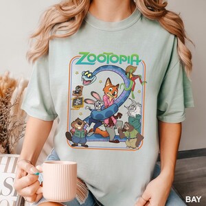 Puede incluir: Camiseta verde claro con la palabra "ZOOTOPIA" en naranja y un dibujo animado de personajes de la pel&iacute;cula. El dibujo animado incluye un zorro, un conejo y otros personajes animales. La camiseta es de cuello redondo.