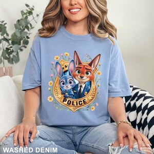 May include: Light blue t-shirt with a graphic of a bunny and fox in police uniforms inside a gold badge with the word "POLICE". The shirt has the words "WASHED DENIM" at the bottom.