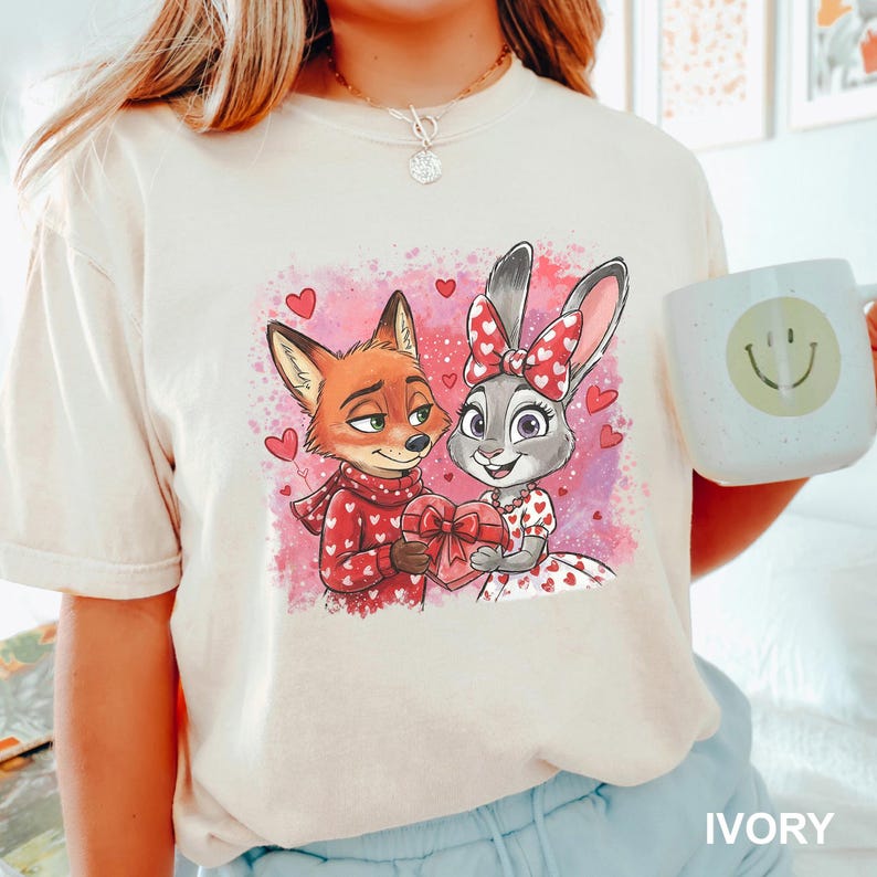 May include: Ivory t-shirt with a cartoon fox and rabbit couple holding a heart-shaped gift. The fox wears a red scarf and sweater with white hearts. The rabbit has a red bow and dress with white hearts. A mug with a smiley face is visible.