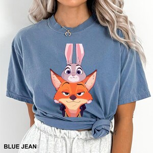 Puede incluir: Camiseta azul vaquero con una ilustraci&oacute;n de dibujos animados de Judy Hopps y Nick Wilde de Zootopia. El texto "BLUE JEAN" est&aacute; impreso en la parte inferior de la camiseta.