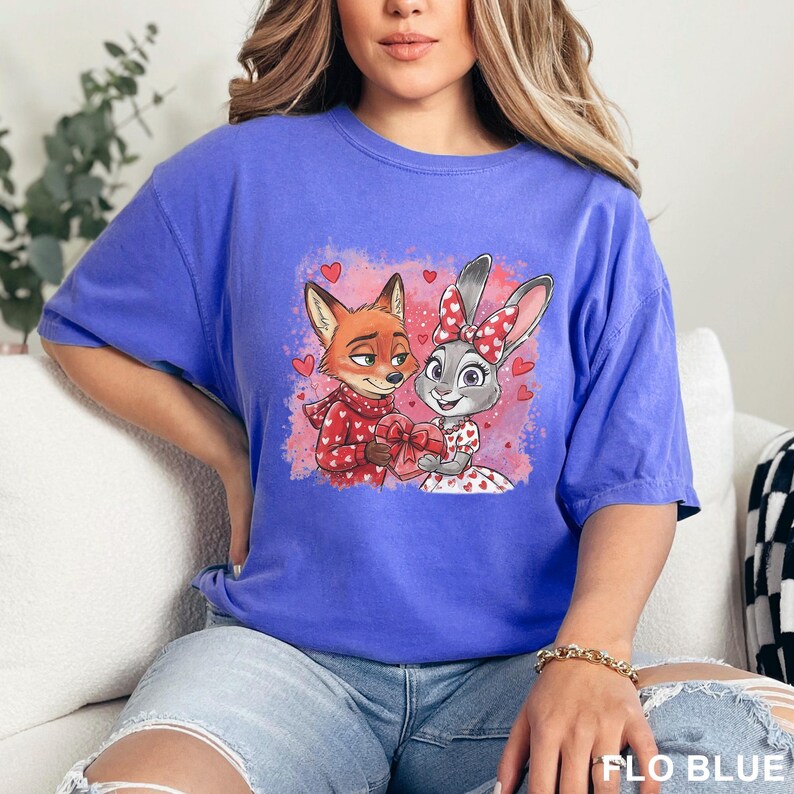 May include: A blue t-shirt with a cartoon image of a fox and a rabbit, both holding a heart-shaped gift. The fox is wearing a red scarf, and the rabbit has a bow. The text "FLO BLUE" is visible at the bottom right.