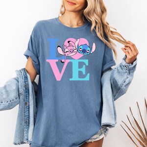 May include: Blue t-shirt featuring the word "LOVE" in pink and blue block letters. The "O" is a pink heart with cartoon characters. The shirt is a solid blue colour.