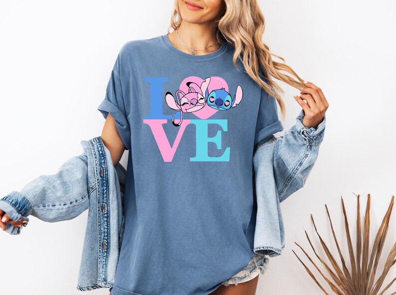 May include: Blue t-shirt featuring the word "LOVE" in pink and blue block letters. The "O" is a pink heart with cartoon characters. The shirt is a solid blue colour.
