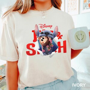 Puede incluir: Camiseta de color marfil con un gr&aacute;fico de Stitch de Disney rompiendo un efecto de papel rasgado, sosteniendo un personaje. La palabra "Disney" est&aacute; encima del gr&aacute;fico, con "1 & Stitch" en rojo. En la imagen se sostiene una taza blanca con una carita sonriente.