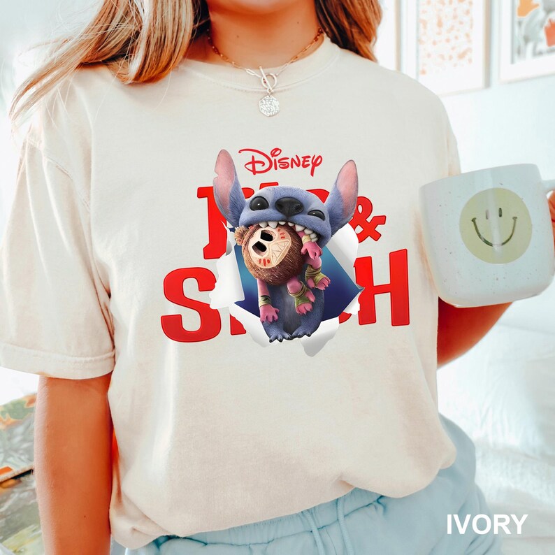 Puede incluir: Camiseta de color marfil con un gr&aacute;fico de Stitch de Disney rompiendo un efecto de papel rasgado, sosteniendo un personaje. La palabra "Disney" est&aacute; encima del gr&aacute;fico, con "1 & Stitch" en rojo. En la imagen se sostiene una taza blanca con una carita sonriente.