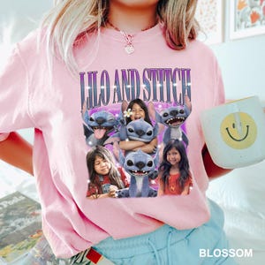 May include: A light pink sweatshirt featuring a graphic of the characters Lilo and Stitch. The graphic includes the text "Lilo and Stitch" in a purple font. A white mug with a yellow smiley face is held in the hand.