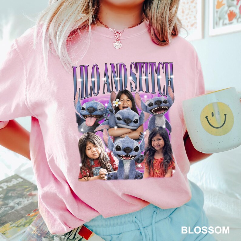 May include: A light pink sweatshirt featuring a graphic of the characters Lilo and Stitch. The graphic includes the text "Lilo and Stitch" in a purple font. A white mug with a yellow smiley face is held in the hand.