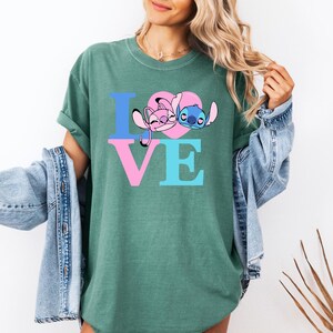 May include: Green t-shirt with the word "LOVE" in blue and pink letters. The "O" is a pink heart with cartoon characters. The shirt is worn with a denim jacket.