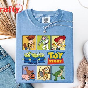 Pu&ograve; includere: Una maglietta blu chiaro con una grafica di personaggi del film Toy Story. I personaggi includono Woody, Buzz Lightyear, Jessie e i tre piccoli alieni verdi. Il testo "Toy Story" &egrave; stampato in lettere bianche su uno sfondo rosso.