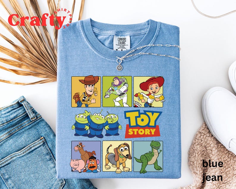 Pu&ograve; includere: Una maglietta blu chiaro con una grafica di personaggi del film Toy Story. I personaggi includono Woody, Buzz Lightyear, Jessie e i tre piccoli alieni verdi. Il testo "Toy Story" &egrave; stampato in lettere bianche su uno sfondo rosso.