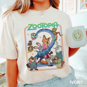 Puede incluir: Camiseta de color marfil con un gr&aacute;fico colorido de la pel&iacute;cula Zootopia. El dise&ntilde;o incluye el t&iacute;tulo "Zootopia" en verde y azul, junto con varios personajes de animales. En la imagen se sostiene una taza blanca con una carita sonriente.