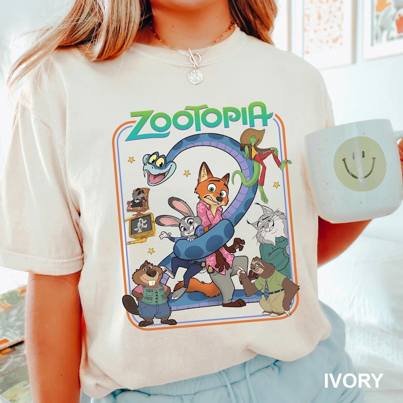Puede incluir: Camiseta de color marfil con un gr&aacute;fico colorido de la pel&iacute;cula Zootopia. El dise&ntilde;o incluye el t&iacute;tulo "Zootopia" en verde y azul, junto con varios personajes de animales. En la imagen se sostiene una taza blanca con una carita sonriente.
