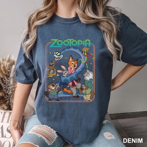 Puede incluir: Camiseta azul denim con un gr&aacute;fico colorido de la pel&iacute;cula Zootopia. El dise&ntilde;o incluye el t&iacute;tulo de la pel&iacute;cula y varios personajes, como un zorro, un conejo y un perezoso. La palabra "DENIM" est&aacute; impresa en la parte inferior derecha.
