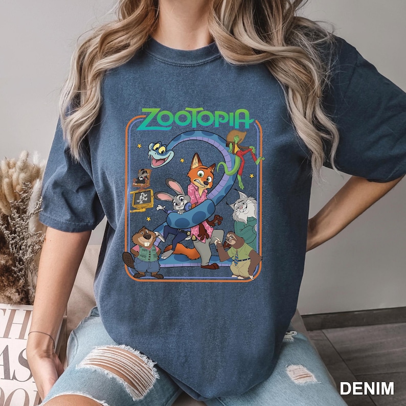 Puede incluir: Camiseta azul denim con un gr&aacute;fico colorido de la pel&iacute;cula Zootopia. El dise&ntilde;o incluye el t&iacute;tulo de la pel&iacute;cula y varios personajes, como un zorro, un conejo y un perezoso. La palabra "DENIM" est&aacute; impresa en la parte inferior derecha.