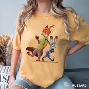 Może przedstawiać: Musztardowa koszulka z kresk&oacute;wkową ilustracją Nicka Wilde'a i Judy Hopps z filmu Zwierzogr&oacute;d. Nick, lis, ma na sobie zieloną koszulę, a Judy, kr&oacute;lik, jest w niebieskim mundurze. Słowo "MUSTARD" jest wydrukowane w prawym dolnym rogu.