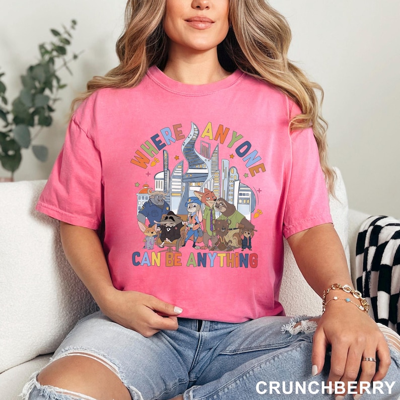 May include: A pink t-shirt with the text "WHERE ANYONE CAN BE ANYTHING" in colorful letters. The graphic features cartoon characters and a cityscape. The shirt is a crew neck and has short sleeves.