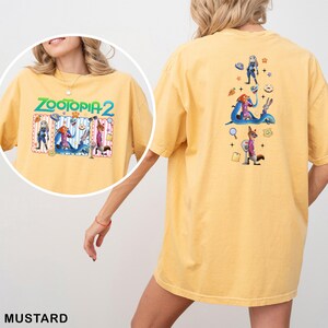 May include: Mustard-colored t-shirt featuring the text "ZOOTOPIA 2" with cartoon characters. The back of the shirt displays additional Zootopia characters and icons. The shirt is a comfortable, casual style.