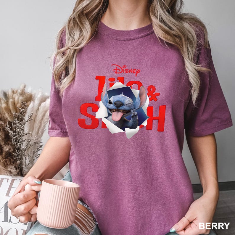 May include: Berry-colored t-shirt featuring a graphic of Stitch from Disney's Lilo & Stitch. The design shows Stitch bursting through a torn paper effect, with the movie title in red. The shirt is a short-sleeved, casual wear item.