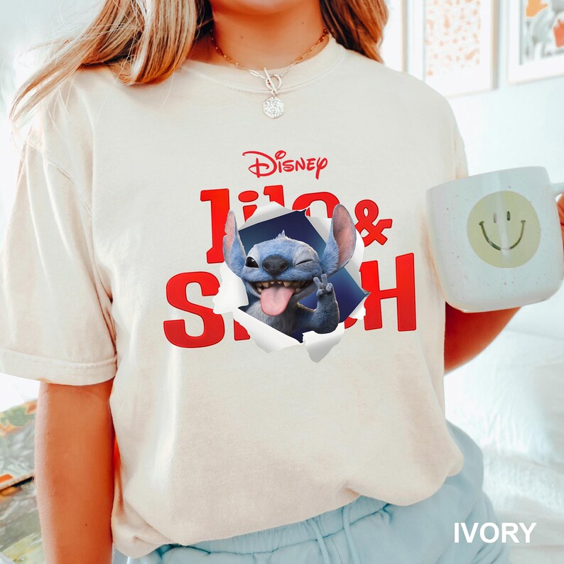 May include: Ivory-colored t-shirt featuring the Disney logo and the words "Lilo & Stitch" in red. The graphic shows Stitch popping out of a torn hole, sticking out his tongue and making a peace sign. A white mug with a smiley face is also visible.