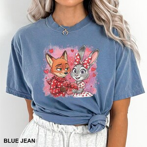May include: Blue jean coloured t-shirt featuring a cartoon fox and bunny holding a heart-shaped gift. The fox wears a red sweater with white hearts, and the bunny wears a dress with heart patterns. The text "BLUE JEAN" is at the bottom.