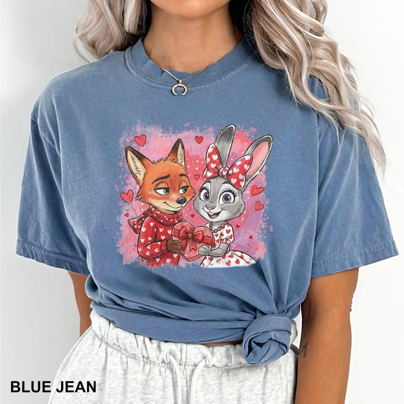 May include: Blue jean coloured t-shirt featuring a cartoon fox and bunny holding a heart-shaped gift. The fox wears a red sweater with white hearts, and the bunny wears a dress with heart patterns. The text "BLUE JEAN" is at the bottom.