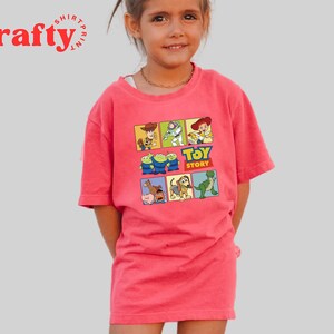 Pu&ograve; includere: Una maglietta rosa con una grafica colorata di personaggi del film Toy Story. La grafica include Woody, Buzz Lightyear, Rex e altri personaggi. Il testo "Toy Story" &egrave; stampato in lettere gialle sulla grafica.