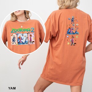 May include: Orange t-shirt featuring the text "ZOOTOPIA 2" with cartoon characters. The front of the shirt has a circular graphic with characters. The back of the shirt has a graphic with characters and other elements.