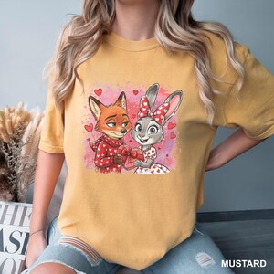 May include: Mustard-coloured t-shirt with a cartoon image of a fox and a rabbit holding a heart-shaped gift box, surrounded by hearts. The fox wears a red scarf, and the rabbit has a red bow. The word "MUSTARD" is printed at the bottom right.