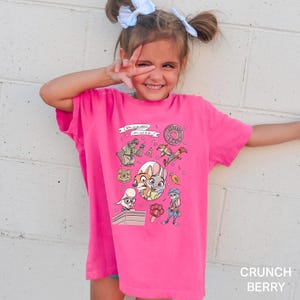 Puede incluir: Una camiseta rosa brillante con personajes de dibujos animados y el texto "THE BIG DONUT". La camiseta tiene cuello redondo y mangas cortas. Las palabras "Crunch Berry" est&aacute;n impresas en la parte inferior derecha.