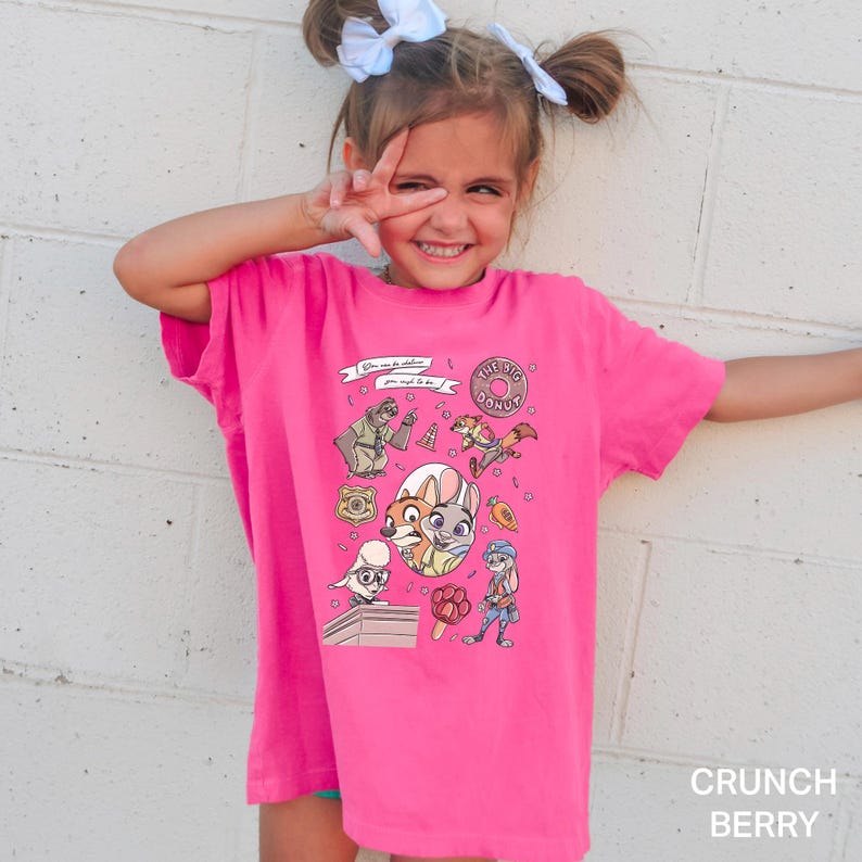 Puede incluir: Una camiseta rosa brillante con personajes de dibujos animados y el texto "THE BIG DONUT". La camiseta tiene cuello redondo y mangas cortas. Las palabras "Crunch Berry" est&aacute;n impresas en la parte inferior derecha.