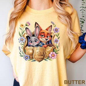 Pu&ograve; includere: T-shirt giallo burro con una grafica colorata di Judy Hopps e Nick Wilde di Zootropolis, incorniciata da fiori e distintivi della polizia. La parola "BUTTER" &egrave; stampata in basso.