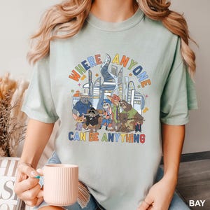 May include: A light green t-shirt featuring a colorful graphic of various cartoon animals and a cityscape. The text "WHERE ANYONE CAN BE ANYTHING" is displayed above the characters in a rainbow font. The shirt is a relaxed fit.