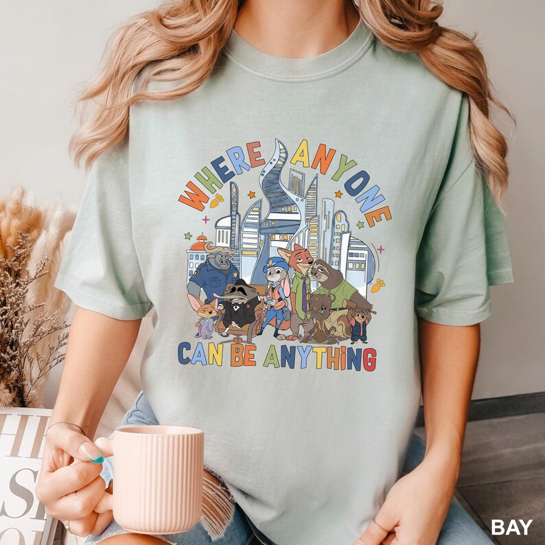 May include: A light green t-shirt featuring a colorful graphic of various cartoon animals and a cityscape. The text "WHERE ANYONE CAN BE ANYTHING" is displayed above the characters in a rainbow font. The shirt is a relaxed fit.