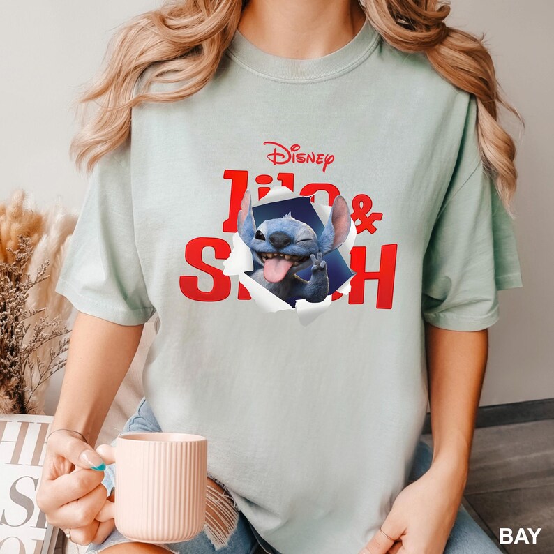 May include: Light green t-shirt featuring the Disney characters Lilo & Stitch. The design shows Stitch popping out of a torn hole, sticking out his tongue. The text "Disney Lilo & Stitch" is in red.