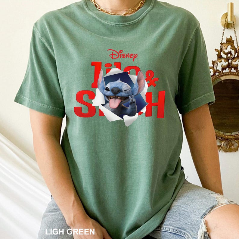 May include: Light green t-shirt featuring the Disney logo and the words "Lilo & Stitch" in red. The graphic shows Stitch popping out of a torn hole. The shirt is a short-sleeved, crew neck style.