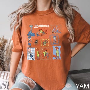 May include: Burnt orange t-shirt featuring a colorful graphic of characters from the animated film "Zootopia." The design includes the film's title and various character illustrations in a grid-like pattern. The shirt is made of a soft material.