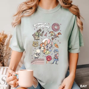Puede incluir: Camiseta verde claro con personajes de dibujos animados de Zootopia. El dise&ntilde;o incluye a Judy Hopps, Nick Wilde y otros personajes, junto con el texto "You can be whatever you wish to be" y "The Big Donut".