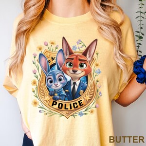 May include: Yellow t-shirt featuring a cartoon image of Judy Hopps and Nick Wilde from Zootopia inside a police badge. The badge is surrounded by flowers and the word "POLICE" is printed at the bottom. The word "BUTTER" is printed at the bottom right.