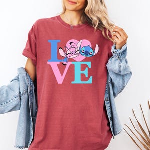 May include: A maroon t-shirt with the word "LOVE" in blue and pink. The "O" is replaced by a pink heart with cartoon characters. The shirt is a casual style.
