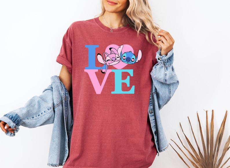 May include: A maroon t-shirt with the word "LOVE" in blue and pink. The "O" is replaced by a pink heart with cartoon characters. The shirt is a casual style.