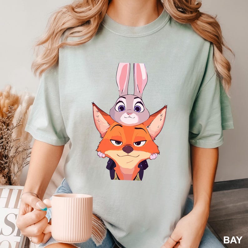 Puede incluir: Camiseta verde claro con una ilustraci&oacute;n de dibujos animados de un zorro y un conejo. El zorro es naranja con una chaqueta negra, y el conejo es gris con orejas rosas. La camiseta es de color liso y tiene un corte holgado.