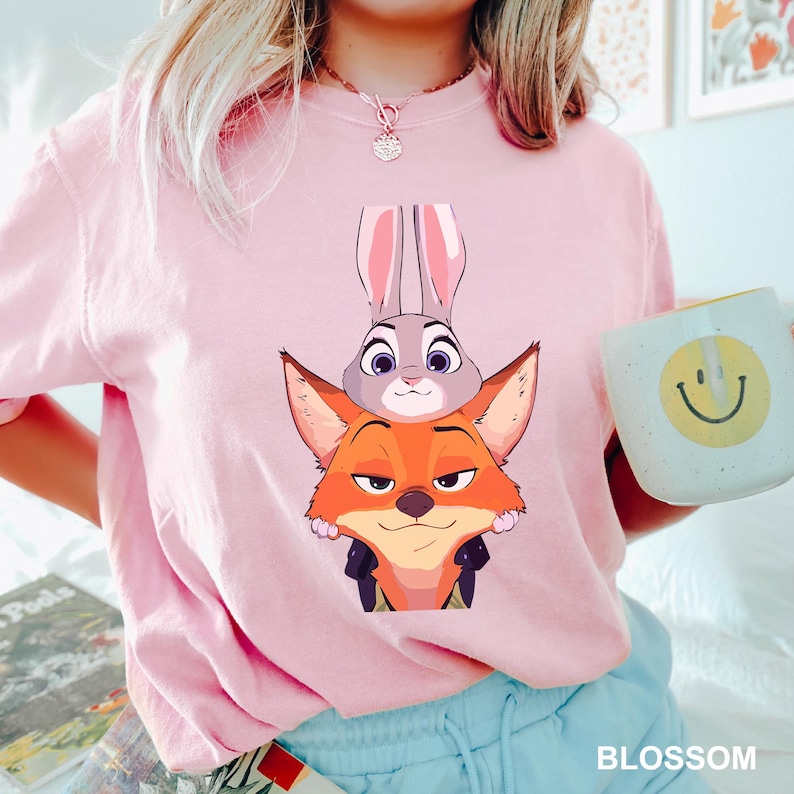 Puede incluir: Camiseta rosa claro con una ilustraci&oacute;n de dibujos animados de Judy Hopps y Nick Wilde de Zootopia. La palabra "BLOSSOM" est&aacute; impresa en la parte inferior. La persona tambi&eacute;n sostiene una taza con una carita sonriente.