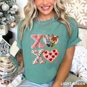 May include: Light green t-shirt with embroidered "XO" design. The "X" letters are pink, one knitted and the other with a black and white polka dot pattern. The "O" is a heart with red hearts. Features a cartoon image of Judy Hopps and Nick Wilde.