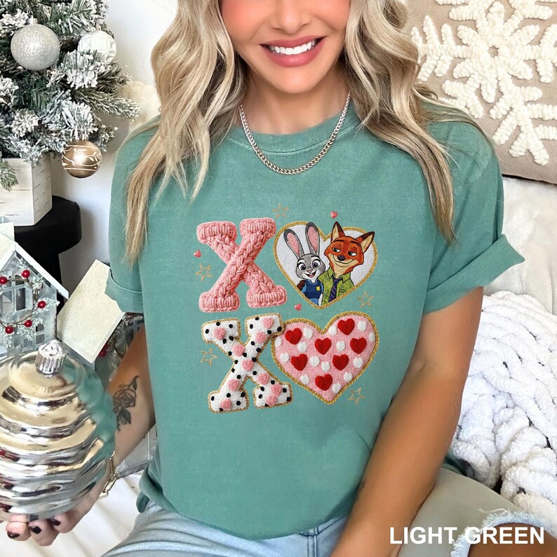 May include: Light green t-shirt with embroidered "XO" design. The "X" letters are pink, one knitted and the other with a black and white polka dot pattern. The "O" is a heart with red hearts. Features a cartoon image of Judy Hopps and Nick Wilde.