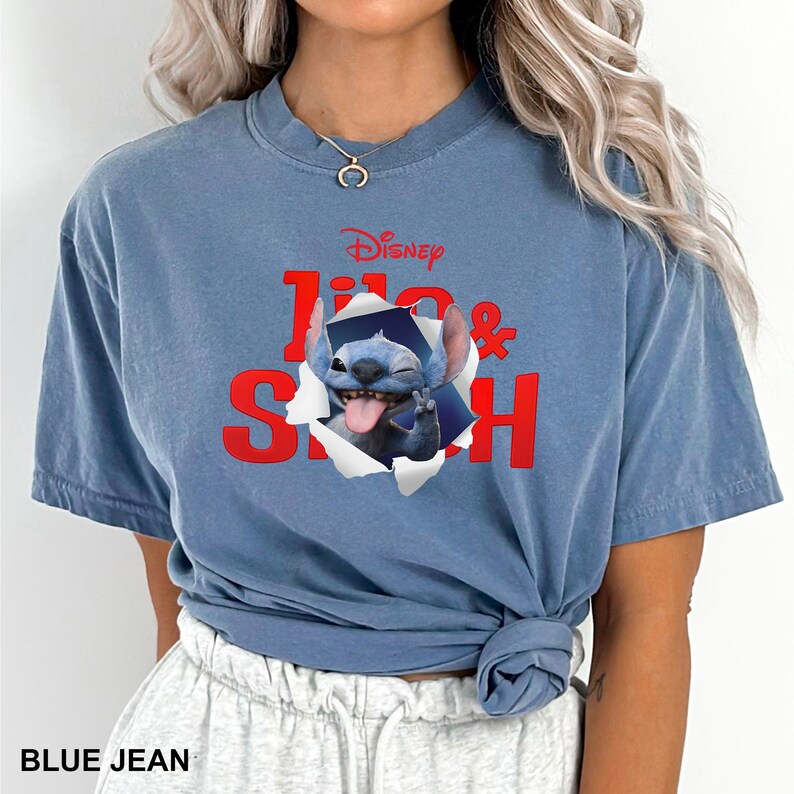 May include: Blue jean-colored t-shirt featuring a graphic of Stitch from Disney's Lilo & Stitch. The design shows Stitch breaking through a torn paper effect, with the Disney logo and the words "Lilo & Stitch" in red. The shirt has a crew neck and short sleeves.
