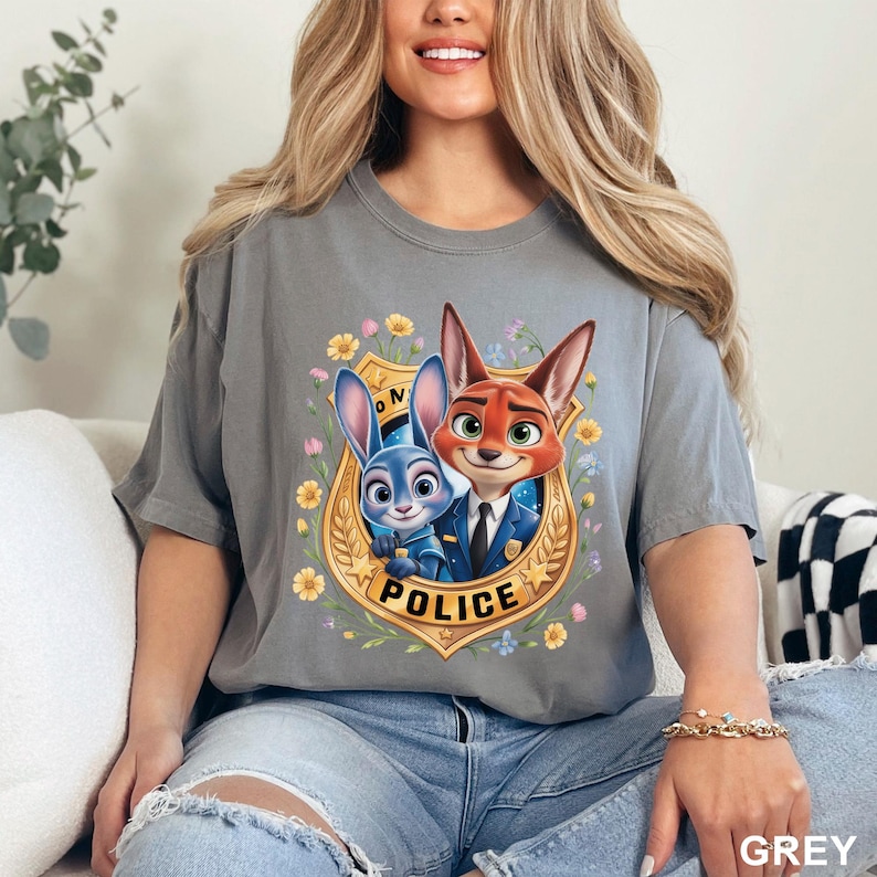 May include: Grey t-shirt featuring a cartoon image of Judy Hopps and Nick Wilde from Zootopia. The characters are inside a police badge with the word "POLICE" and surrounded by flowers. The word "GREY" is in the bottom right corner.