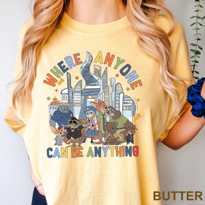 May include: Yellow t-shirt with a colorful graphic featuring cartoon characters and a cityscape. The text on the shirt reads "WHERE ANYONE CAN BE ANYTHING". The shirt is made of a soft material.